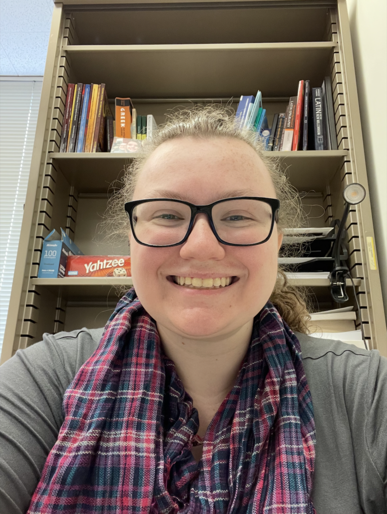 Alumn Amy Vandervelde '21 smiles in a selfie wearing a plaid scarf and gray top, posed in front of a bookshelf.
